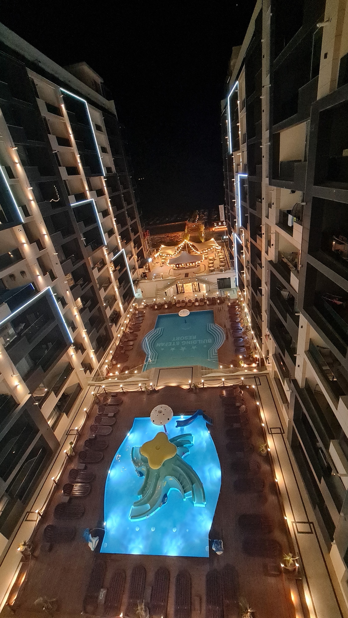 A nighttime view of the apartment complex showcases well-lit pathways leading to a colorful swimming pool area. The pool is highlighted by playful aquatic structures, surrounded by lounge chairs. Soft lights create a welcoming ambiance across the pools and pathways.