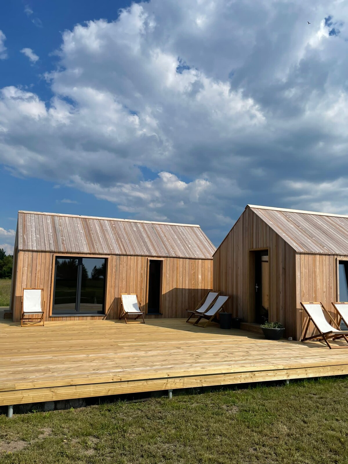 Two wooden structures with sloped roofs are shown on a spacious deck, surrounded by grass. Multiple sun loungers are arranged on the deck, and large glass doors provide access to the inside. The sky is partly cloudy, creating a serene backdrop.