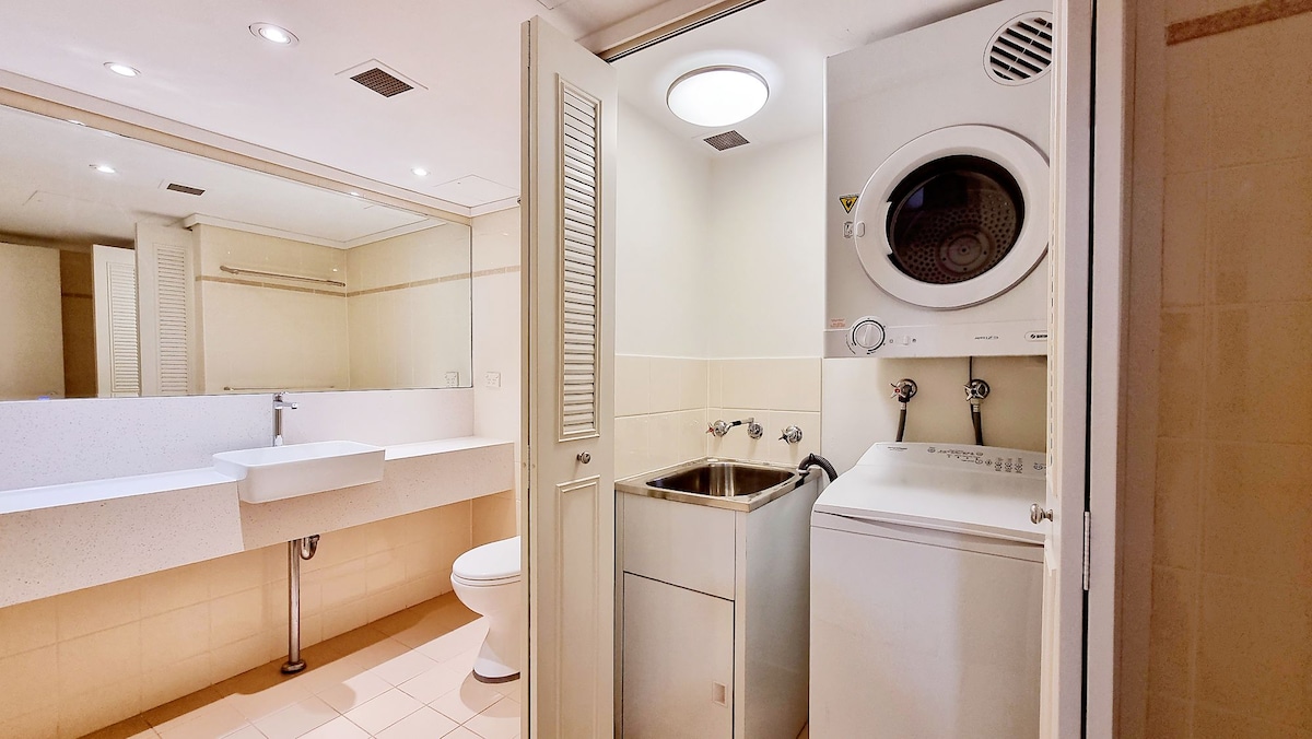 A modern laundry area features a stacked washer and dryer unit, alongside a stainless steel laundry tub. A spacious countertop and wall-mounted mirror enhance functionality. Soft lighting creates an inviting ambiance in this clean and well-organized space.