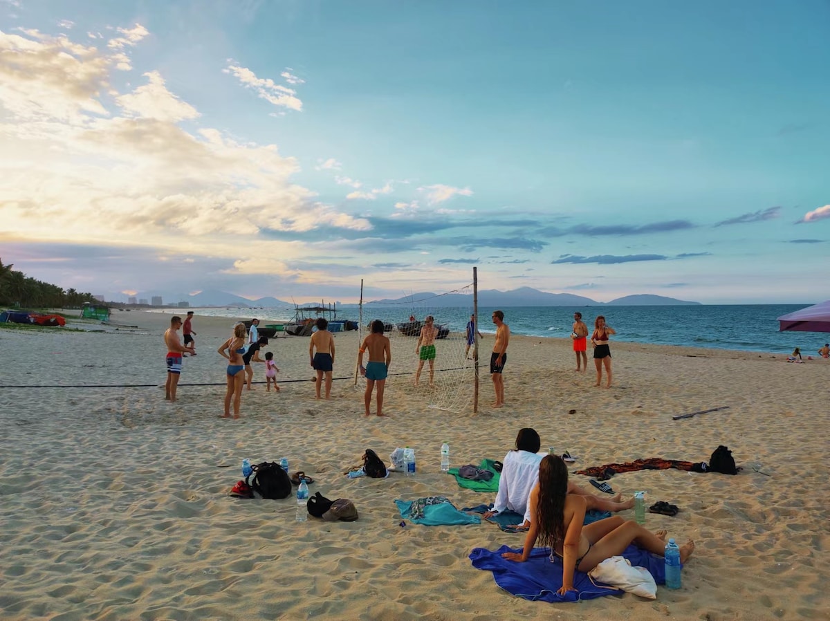 A sandy beach scene is captured at sunset, with groups of people engaged in various activities. Some individuals are playing volleyball near the water's edge, while others relax on towels. The distant horizon features gentle waves and a few boats, creating a tranquil coastal atmosphere.
