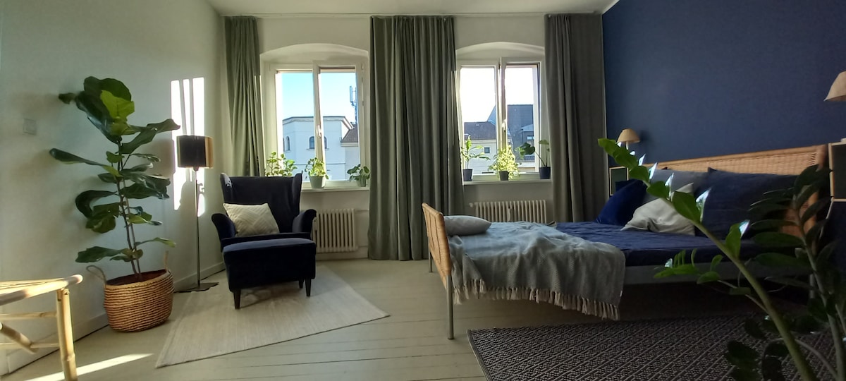 A bright bedroom is featured, showcasing a double bed with a neatly arranged throw. Large windows adorned with sheer curtains allow natural light to fill the space. A comfortable armchair sits beside a floor lamp, while decorative plants add a touch of greenery throughout.