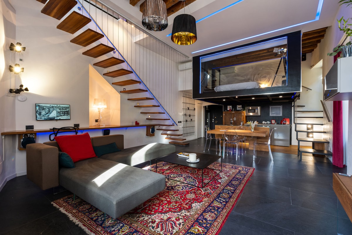 An open living area features a stylish gray sofa adorned with vibrant cushions, a patterned area rug, and wooden stairs leading to an upper loft. Natural light enhances the space, showcasing modern light fixtures and a glass structure above that contains a sleeping area.