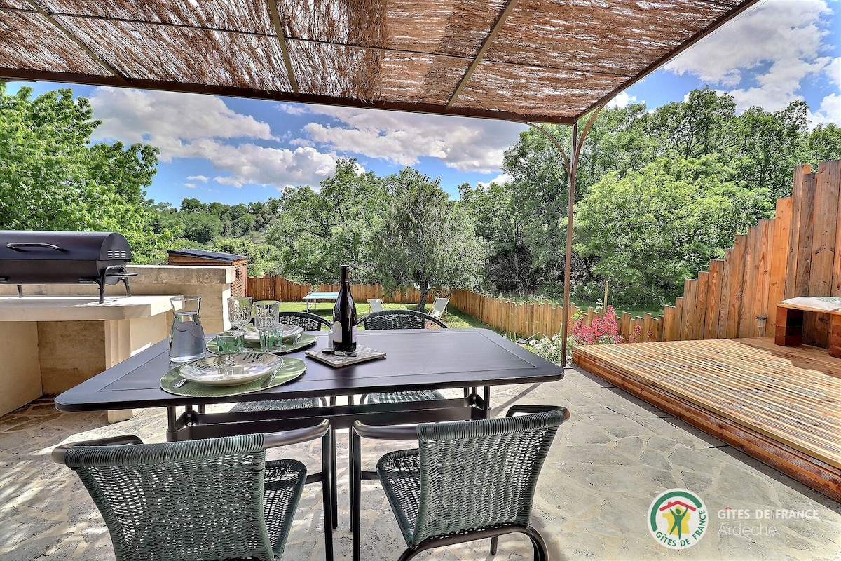 An outdoor dining area is presented with a table set for four, featuring dark wicker chairs and simple dinnerware. A barbecue grill is positioned nearby, and wooden decking is visible in the background. Lush greenery surrounds the space, enhancing the natural setting.