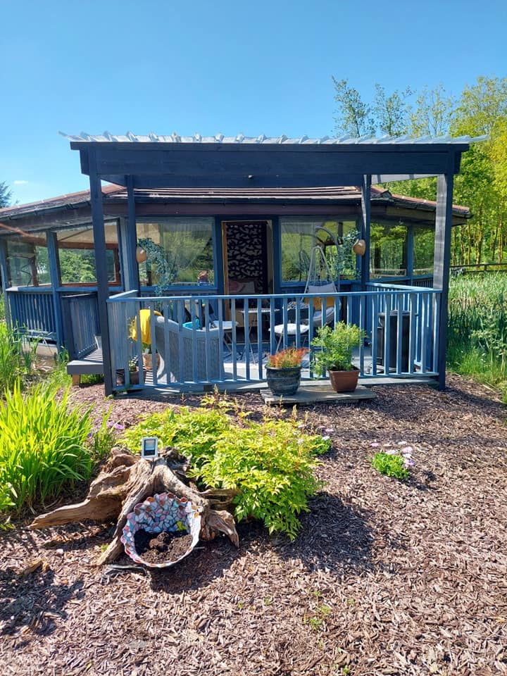 The exterior of the cozy lodge is framed by lush greenery, featuring a blue wooden deck with seating under an overhang. Potted plants are arranged by the entrance, while a weathered log serves as a unique decorative element in the garden.