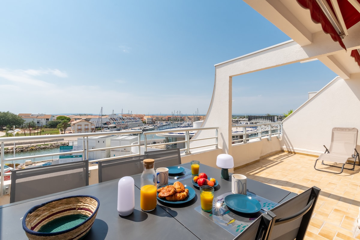 A spacious terrace is set up for outdoor dining, featuring a table with colorful plates, glasses of juice, and an assortment of pastries. The serene view of the harbor and blue sky can be seen in the background, enhancing the relaxed atmosphere.