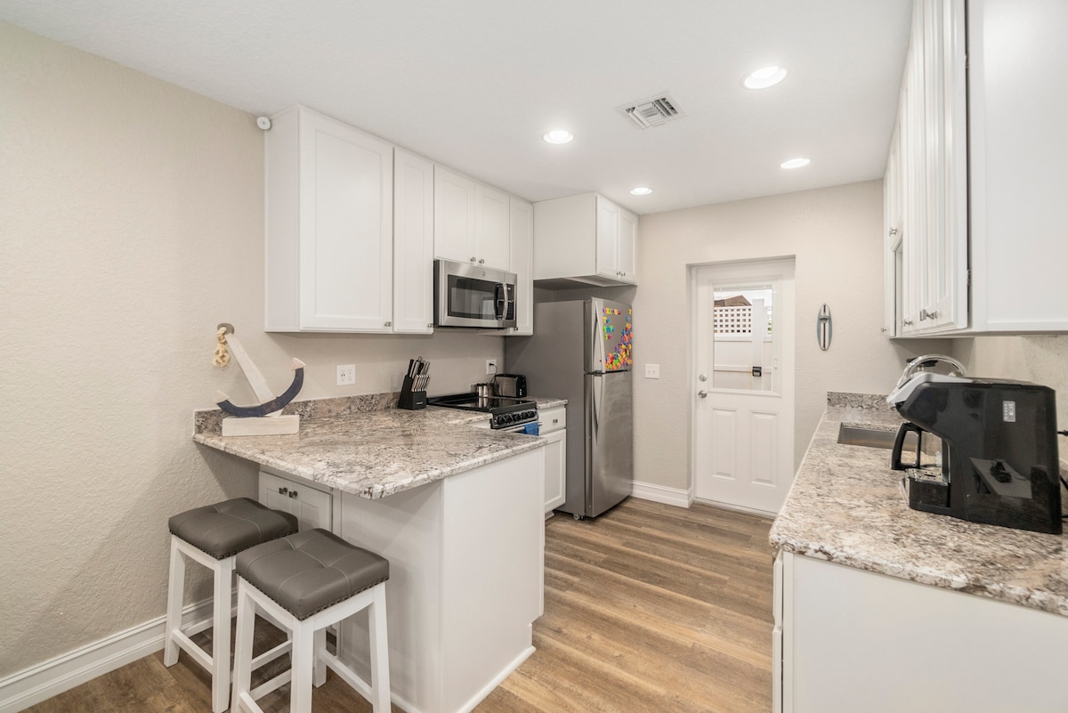 A modern kitchen is presented, featuring white cabinetry and a granite countertop. Stainless steel appliances include a refrigerator and microwave. A small breakfast bar with two stools is situated nearby, offering a functional space for dining. Light fixtures provide ample illumination throughout.