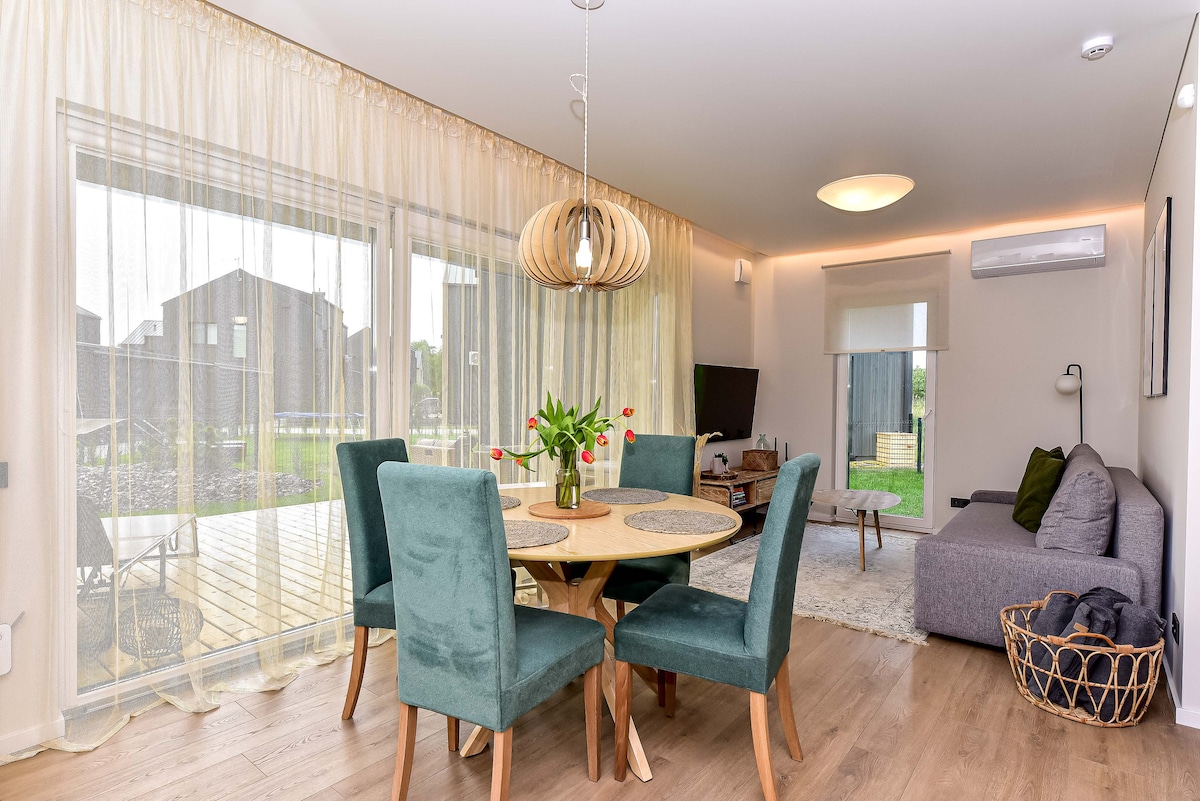 A dining area features a round table surrounded by four upholstered chairs, illuminated by a modern pendant light. A cozy seating area is present, with a gray sofa and a decorative basket nearby. Glass doors open to a private terrace, allowing natural light to fill the space.