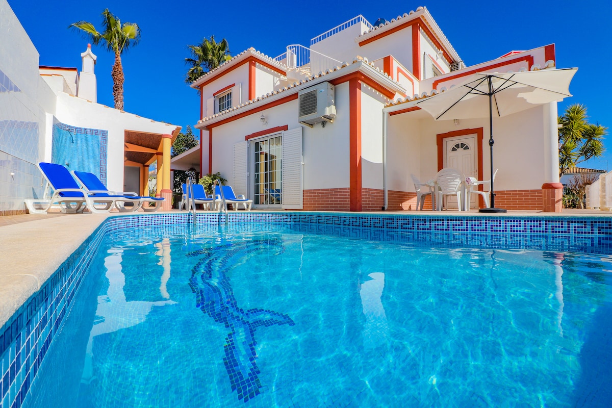 A private pool reflects the bright blue sky, surrounded by sun loungers. The spacious two-story house features a welcoming entrance, with an umbrella providing shade for outdoor seating. Lush greenery is visible, enhancing the tranquil atmosphere of the residential area.