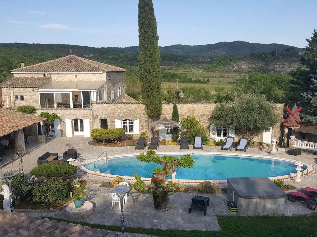 A charming stone residence is set amid lush greenery, featuring a large outdoor swimming pool surrounded by sun loungers. Terraced areas provide various seating options. The backdrop includes rolling hills, enhancing the peaceful atmosphere, while trees offer shaded spots throughout the space.