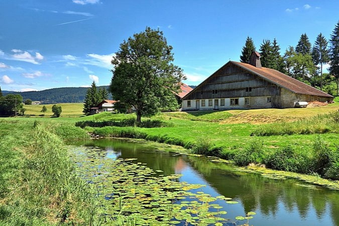 A spacious farm building is nestled beside a gently flowing stream, surrounded by lush green fields. The setting includes large trees and distant hills, with the sky displaying a few scattered clouds and bright sunlight casting a warm glow across the landscape.