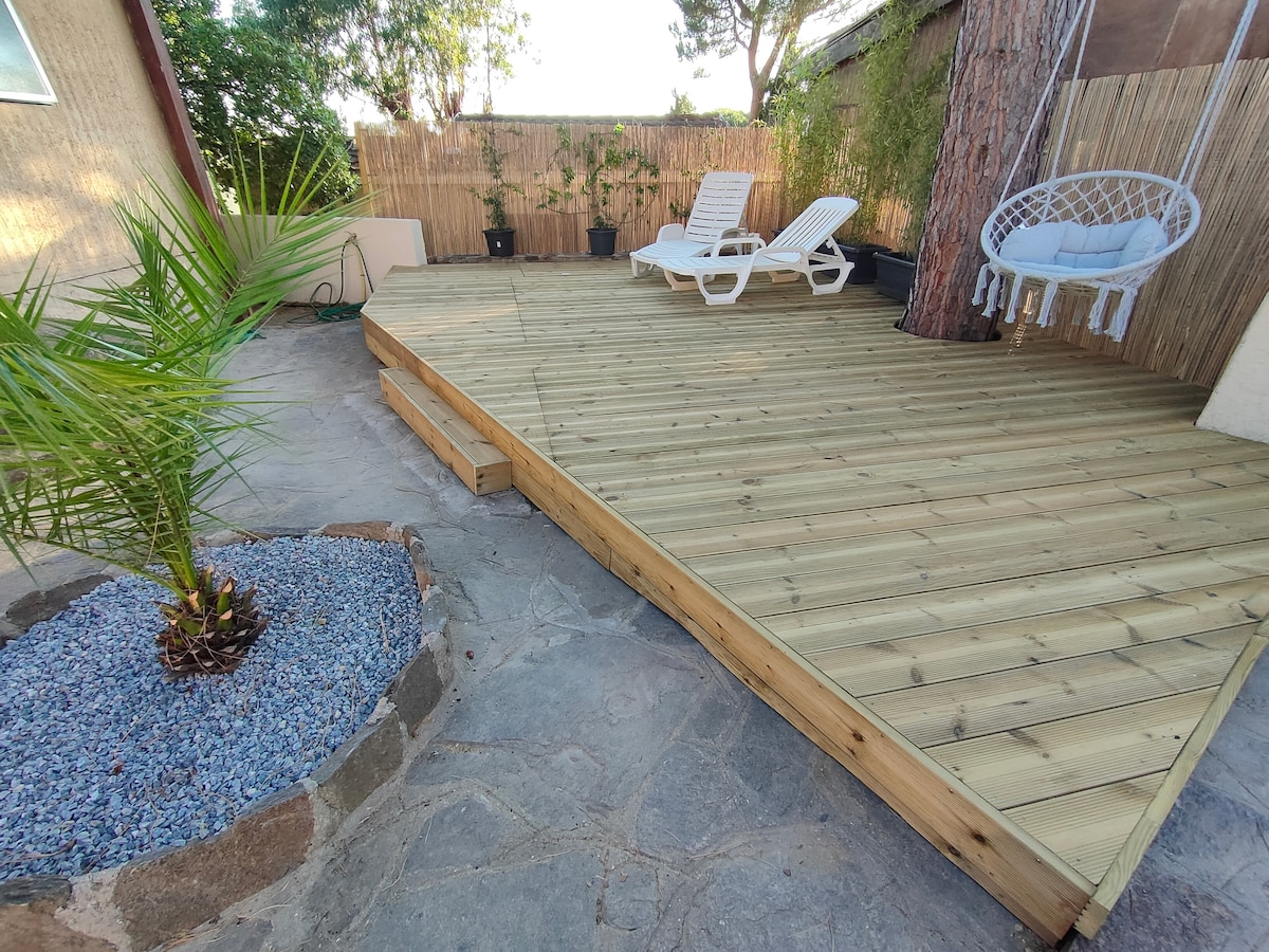 A spacious wooden deck features two lounge chairs and a hanging swing chair. Surrounding greenery includes a small palm and a decorative stone arrangement filled with pebbles, creating a serene outdoor space.