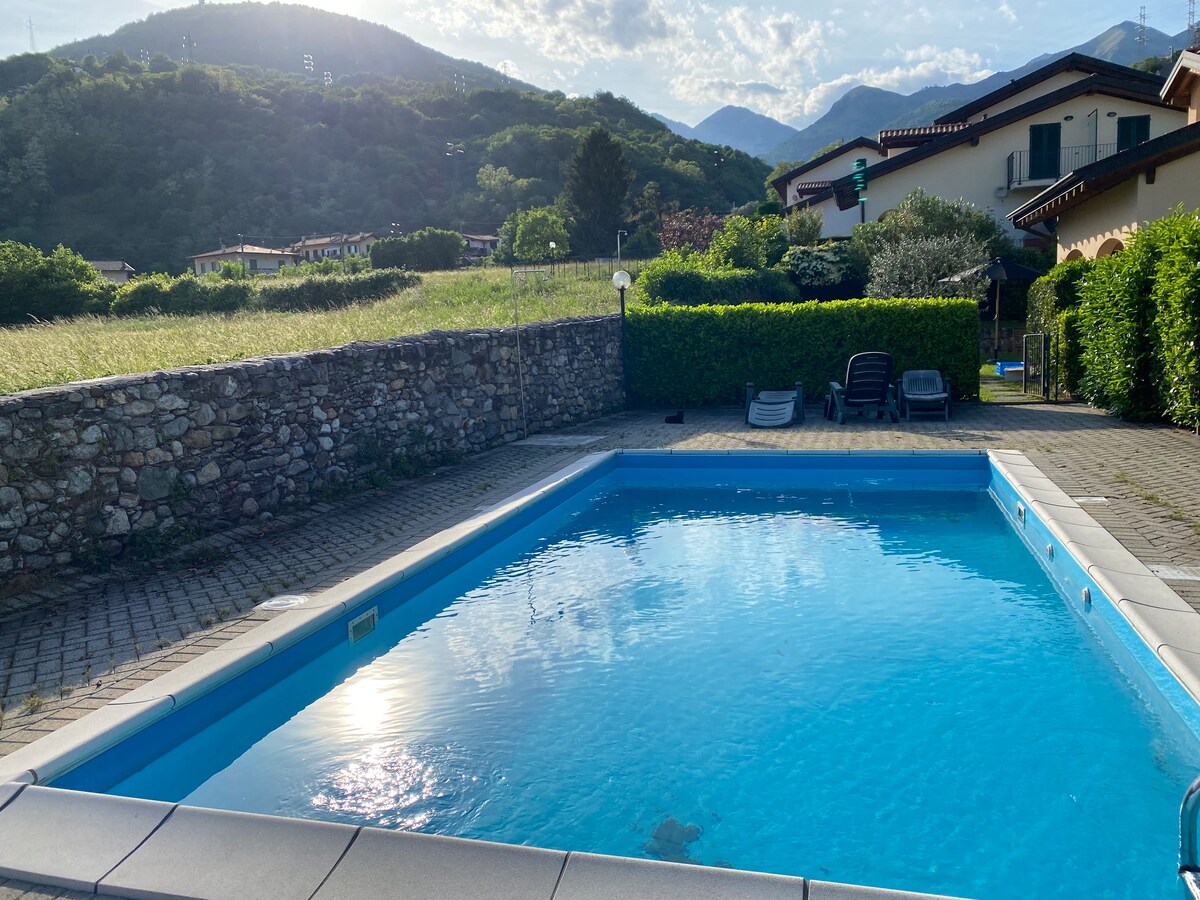 A shared swimming pool is surrounded by neatly trimmed hedges and a stone wall. Sunlight reflects off the clear water, while mountains create a serene backdrop. Lounge chairs are positioned nearby on the paved area, contributing to a tranquil outdoor setting.