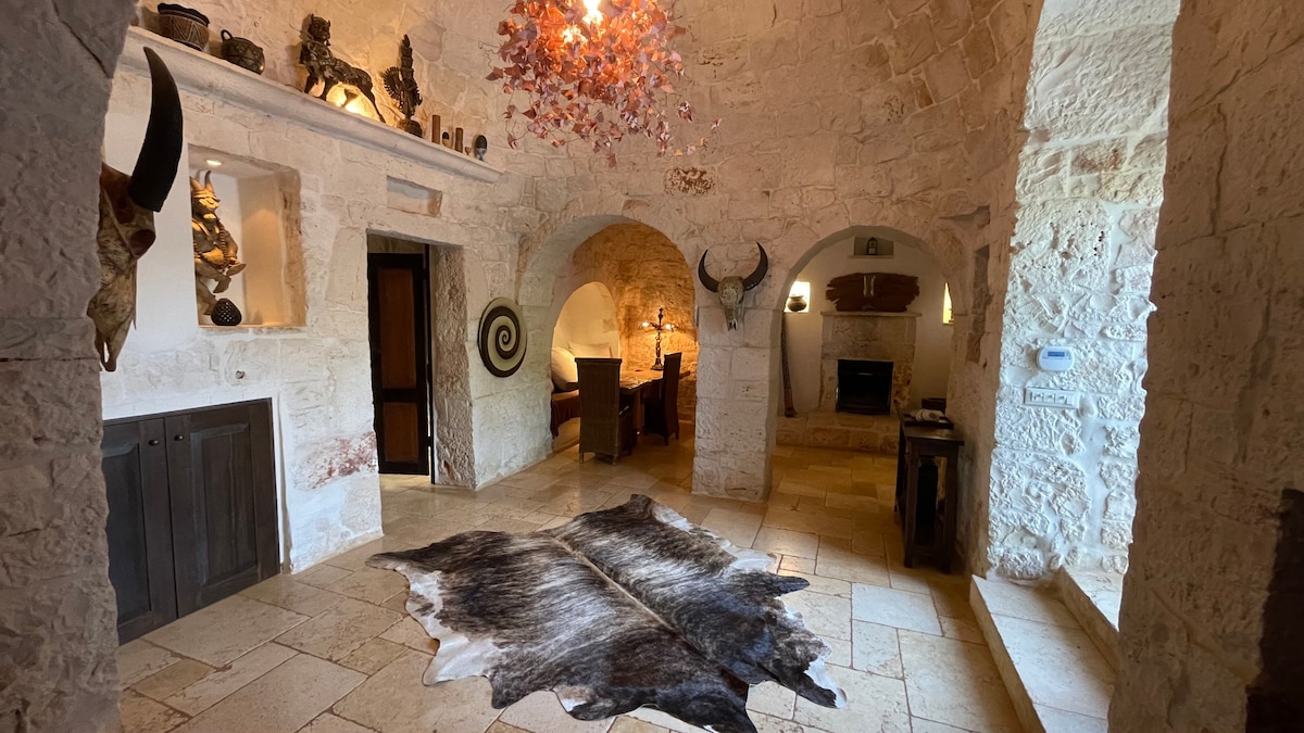 An elegant interior space features rustic stone walls and arched doorways. A large cowhide rug lies on the floor, while decorative items, including sculptures and a fireplace, are positioned on shelves and tabletops. Soft lighting creates a warm ambience throughout the room.