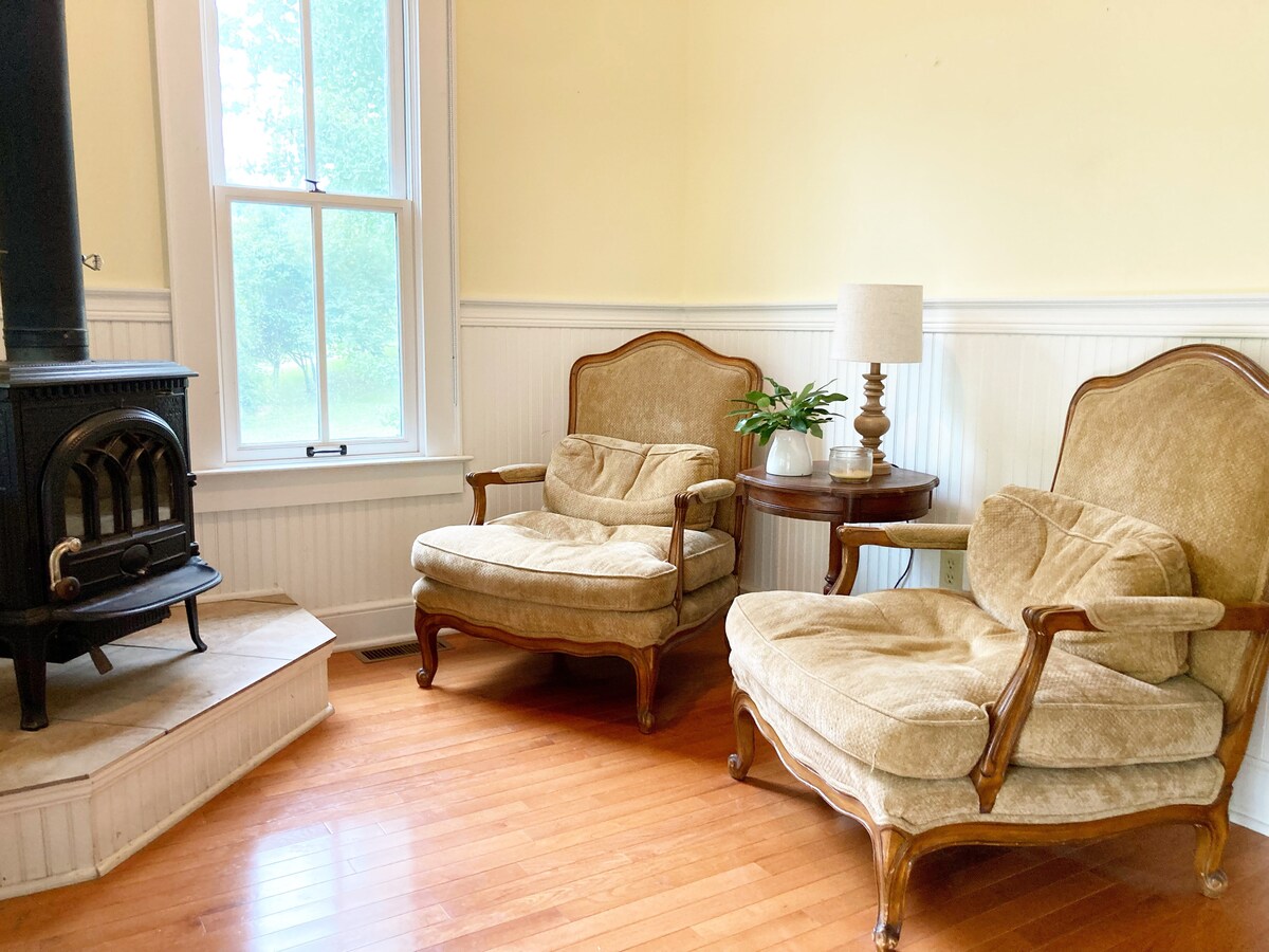 Two vintage armchairs sit invitingly beside a small round table with a lamp and a potted plant. A black wood stove stands nearby against a wall, and natural light streams through a large window, illuminating the warm wooden floors.