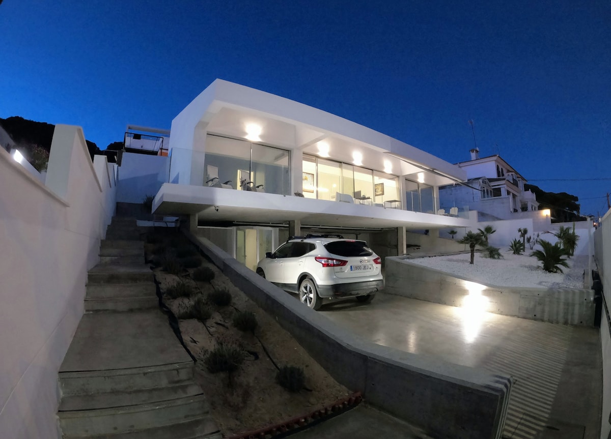 A modern white villa is seen from an elevated angle, positioned on a sloped driveway. The exterior features large glass windows reflecting the evening sky. A silver vehicle is parked on the sleek concrete surface, while a low-maintenance garden with gravel and sparse vegetation occupies the surrounding area.