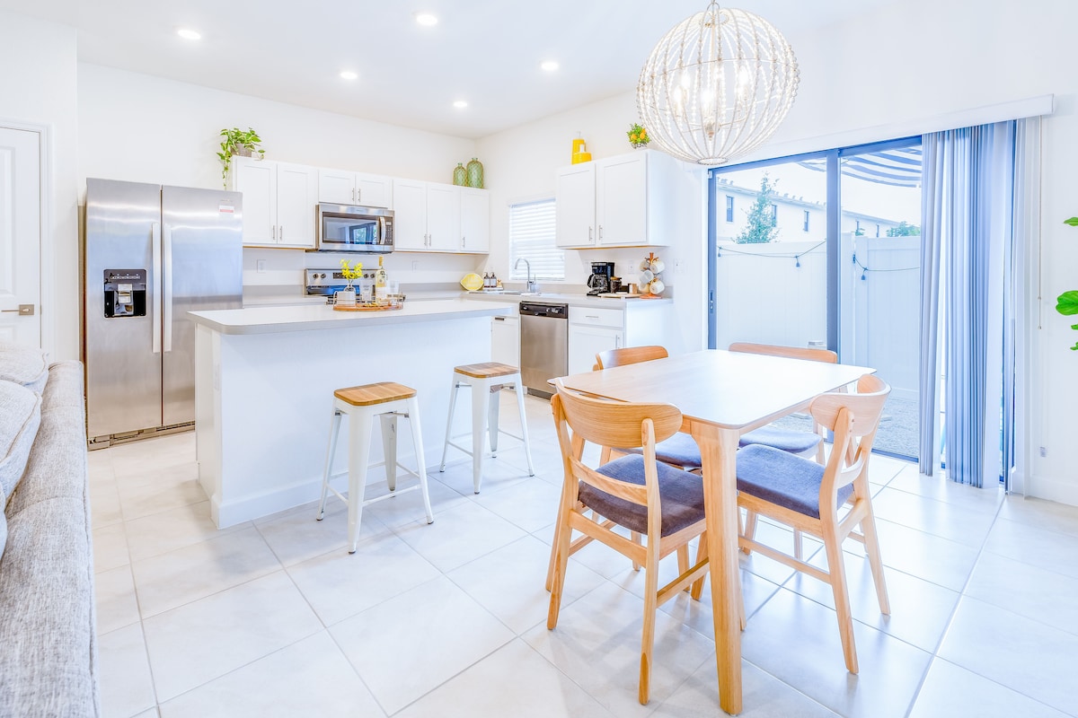 A bright kitchen and dining area features white cabinetry and stainless steel appliances. A central island with bar stools offers additional seating. Natural light enters through sliding glass doors, illuminating a wooden dining table surrounded by four chairs, enhancing the open layout and airy feel.