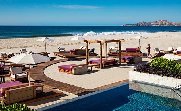 A spacious outdoor lounge area is surrounded by sun beds and umbrellas, inviting relaxation. The coastline is visible in the background, with gentle waves lapping against the sandy beach. A swimming pool features a tranquil water surface, enhancing the serene atmosphere.