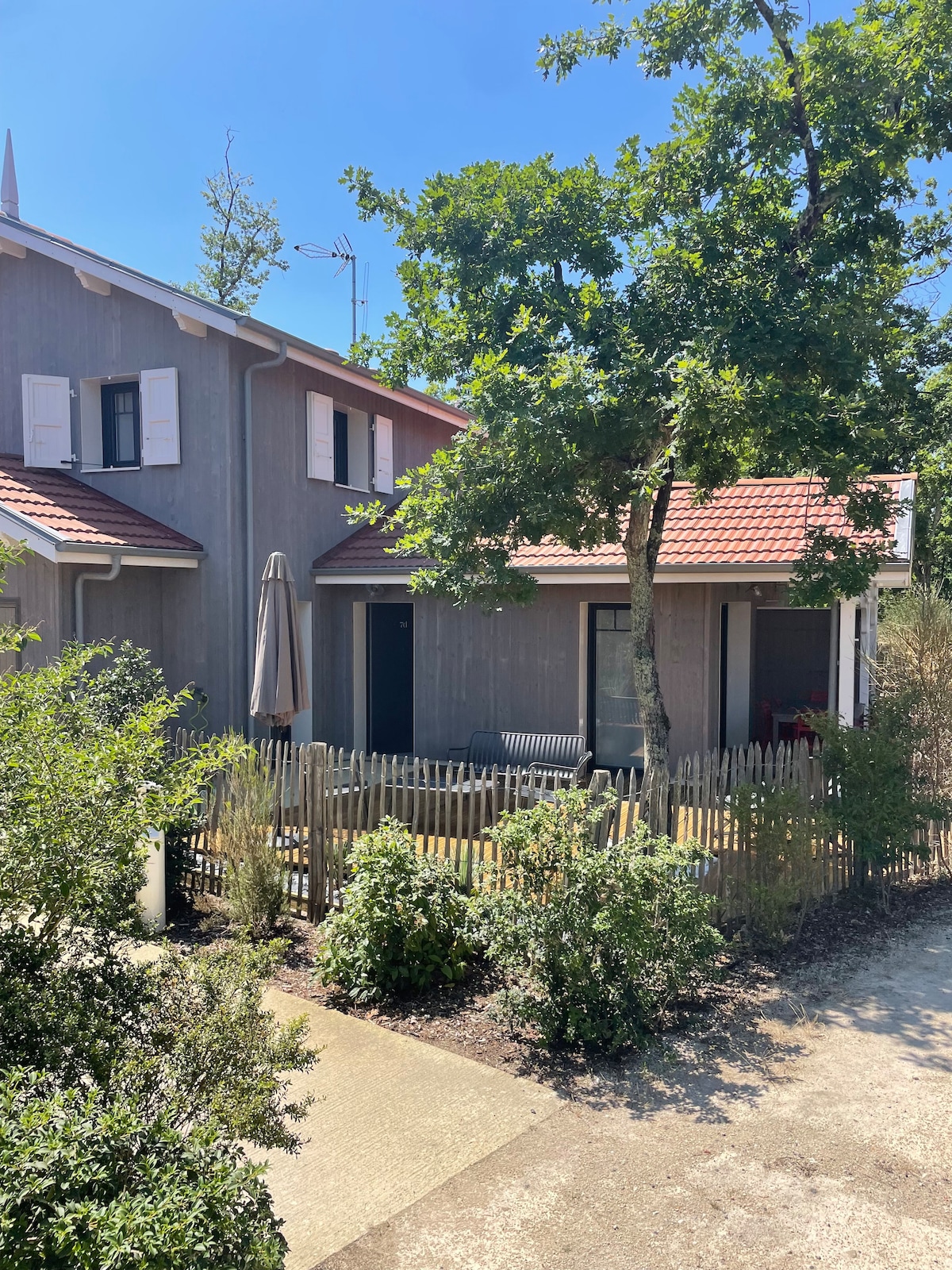 A charming duplex is shown, featuring a wooden exterior and red tiled roof. A small porch with a seating area is visible, surrounded by greenery. A parasol stands next to the building, and a pathway leads to the entrance, enhancing accessibility.