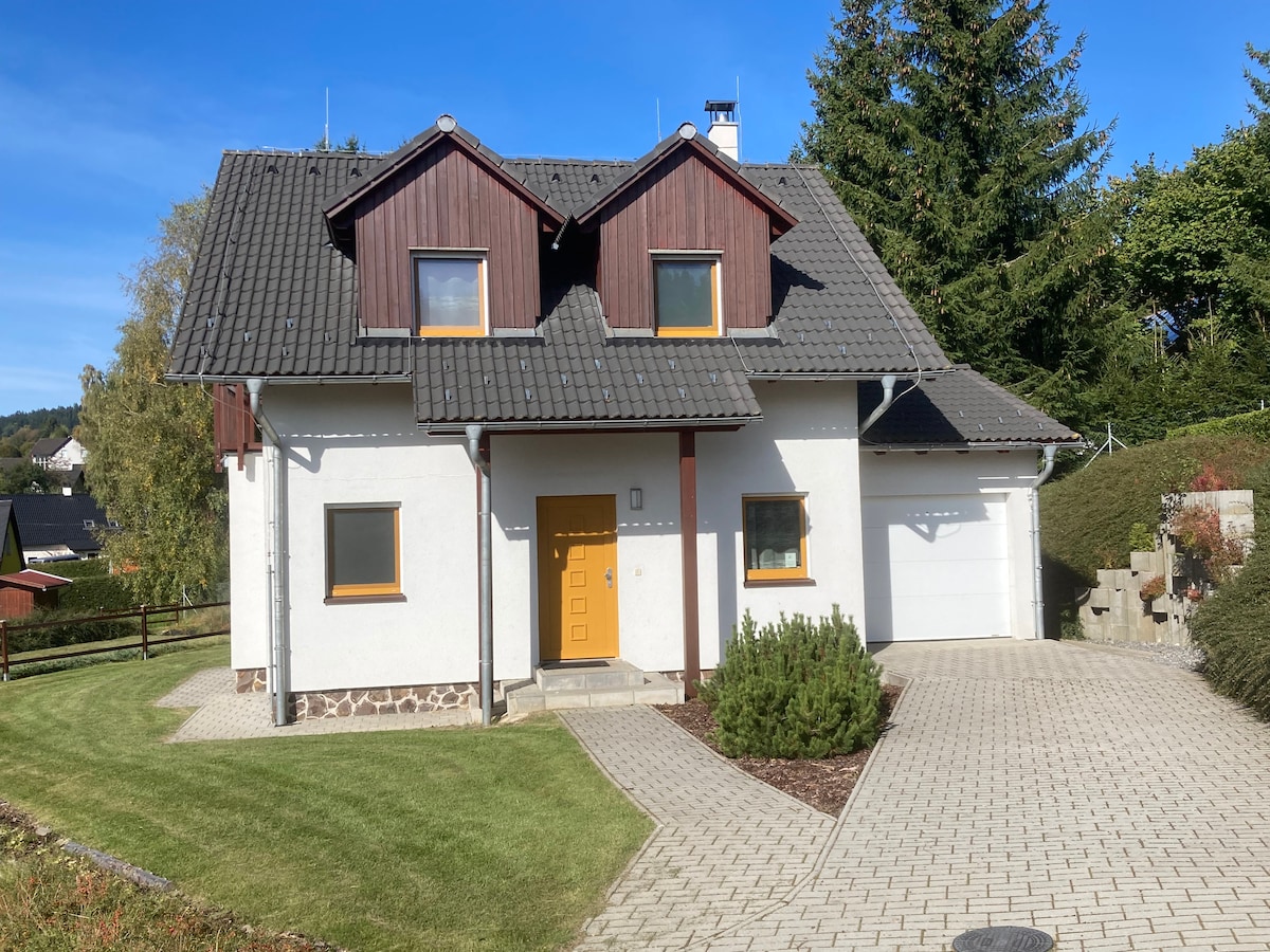 This two-story house features a clean exterior with a combination of white walls and dark wood accents. A vibrant orange door serves as a focal point, while neatly manicured grass and a paved driveway lead to the entrance. Surrounding greenery adds to the natural setting.
