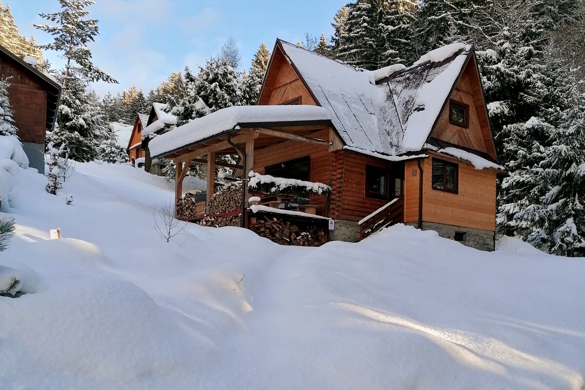 A wooden cabin is nestled among snow-covered trees, featuring a sloped roof and a covered porch. Stacks of firewood are neatly arranged next to the entrance, and the surrounding landscape is blanketed in fresh snow, creating a serene, winter atmosphere.