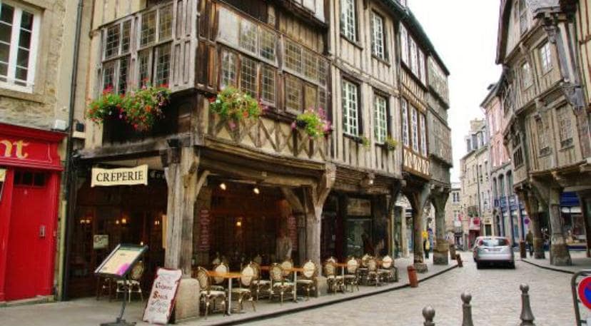An outdoor view of a charming street lined with historic half-timbered buildings. A crêperie with outdoor seating is visible, surrounded by flower planters. The narrow street features cobblestones and is framed by additional colorful buildings, creating a quaint and inviting atmosphere.