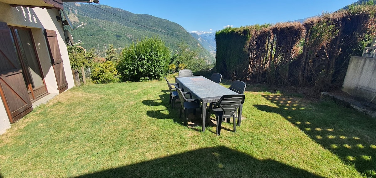 An outdoor space features a large dining table surrounded by several black chairs, situated on a green lawn. Lush greenery is visible in the background, complemented by panoramic views of the mountains and valleys, capturing the essence of a tranquil setting.