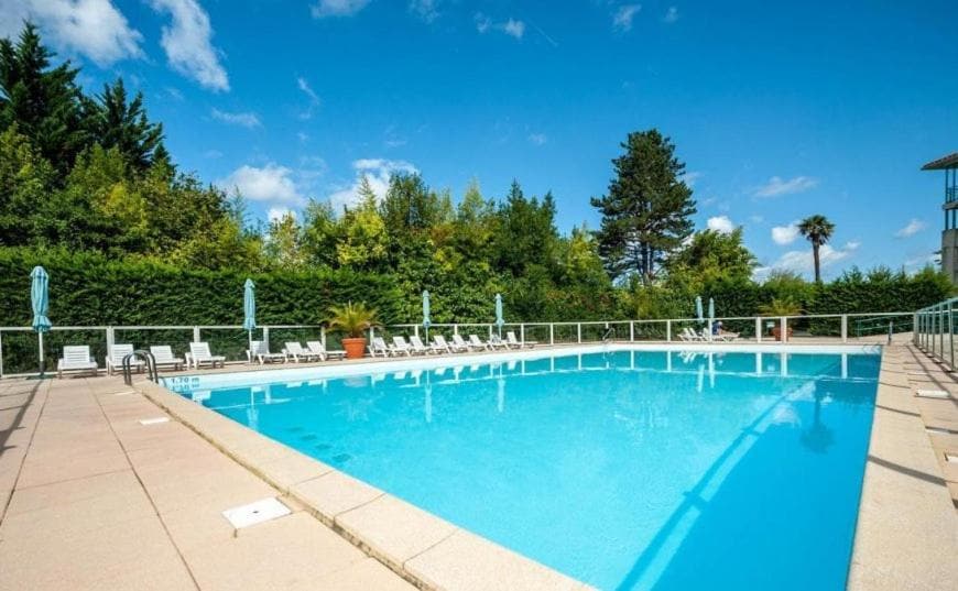 A spacious outdoor pool is surrounded by a sun terrace, featuring several lounge chairs and umbrellas. Lush greenery lines the perimeter, contributing to a serene environment under a blue sky.