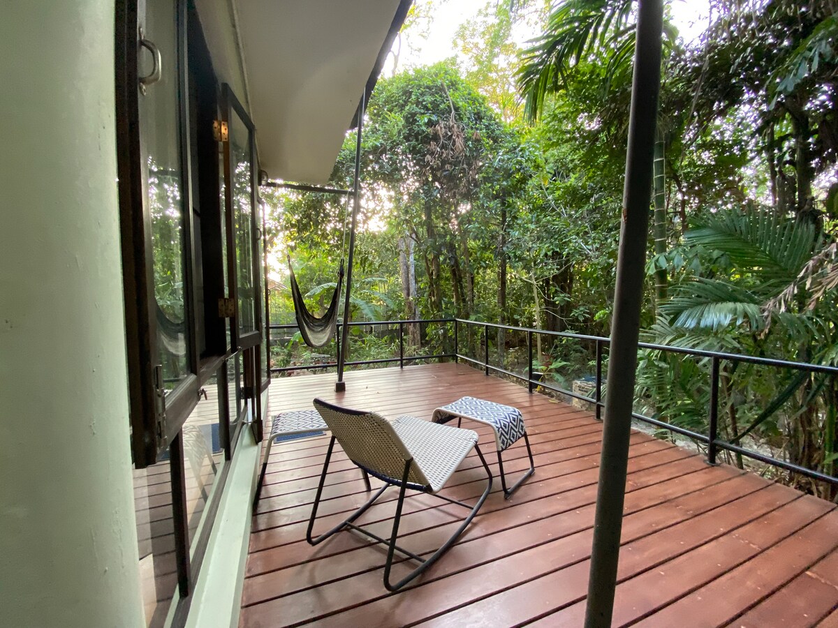A spacious outdoor deck is surrounded by lush greenery, featuring two lounge chairs positioned for relaxation. A hammock is suspended nearby, inviting leisurely moments in nature. The area offers a sense of tranquility, with the soft light filtering through the trees.