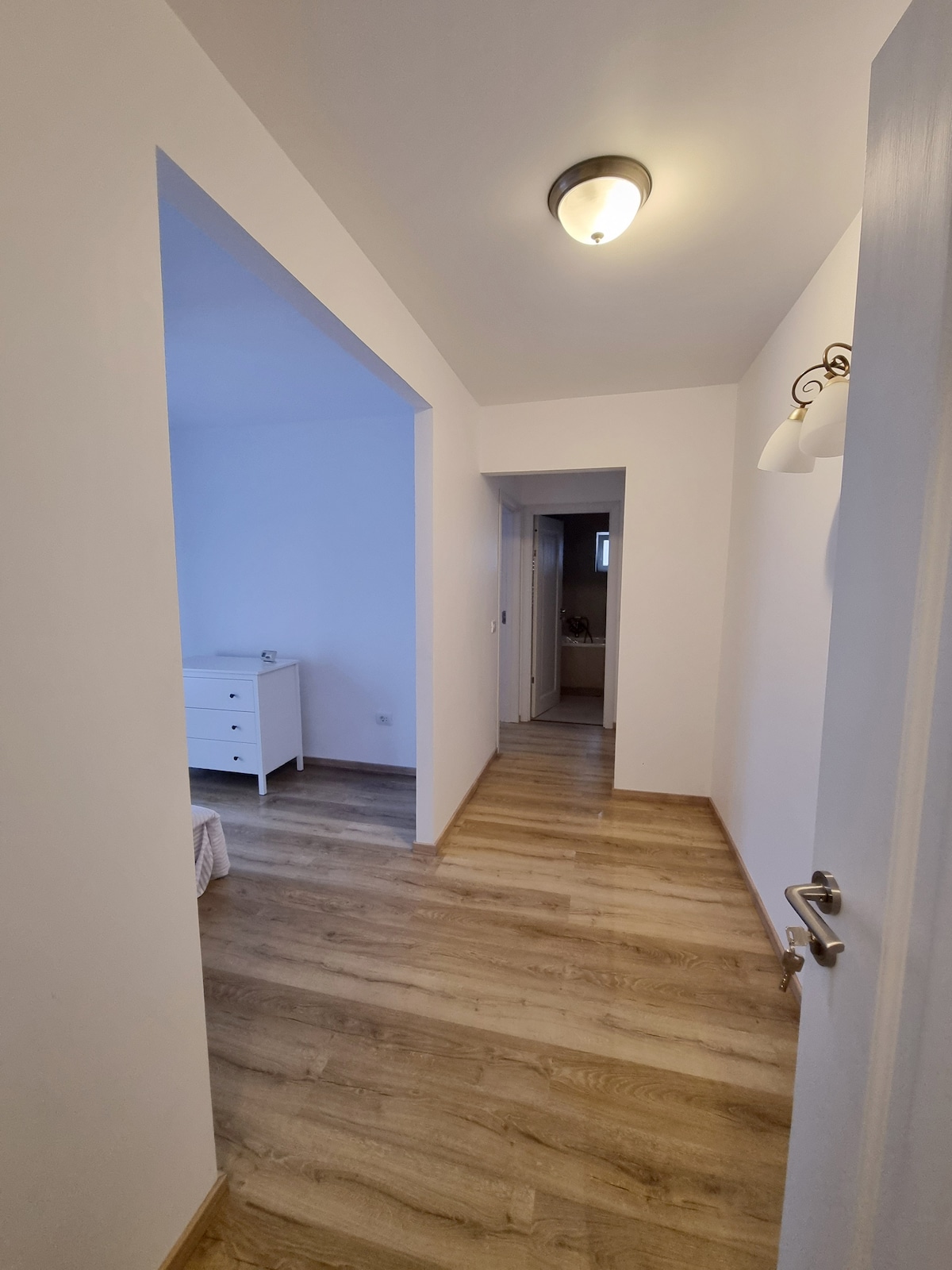 A well-lit hallway showcases smooth wooden flooring and walls painted in a soft white hue. A small white dresser is positioned to the left, while doorways lead to other areas of the apartment. Natural light filters in from the room at the end of the hall.