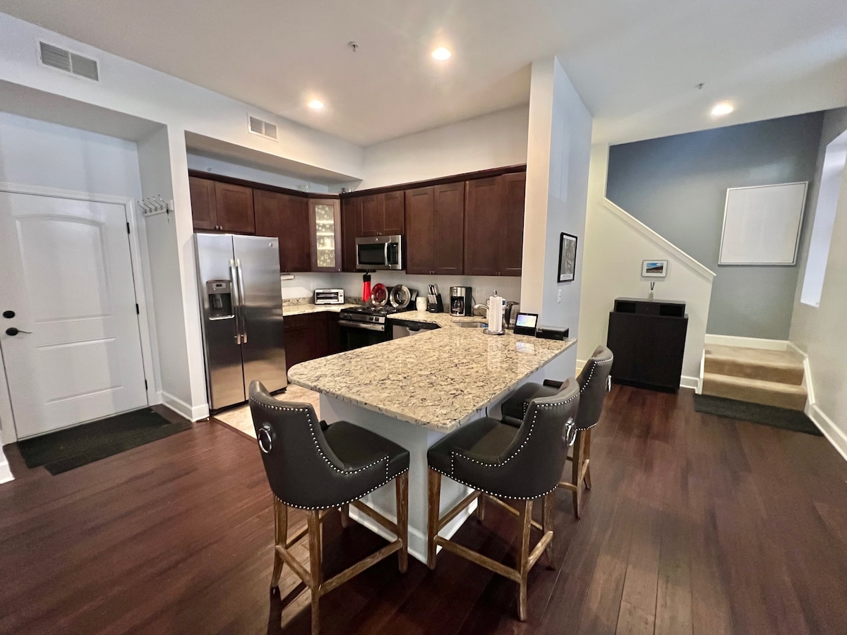 A modern kitchen features dark wooden cabinetry and granite countertops, complemented by stainless steel appliances. An island with seating for three is positioned centrally, while a staircase and a dining area are visible in the background. Natural light illuminates the space, highlighting the rich hardwood floors.