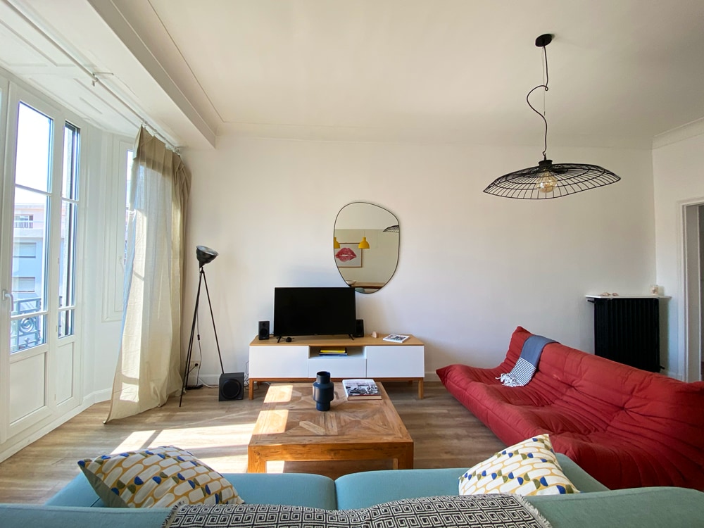 A bright living area features a mix of seating, including a red sofa and a mint green couch, accompanied by a wooden coffee table. A round mirror and a flat-screen TV are also present, with natural light pouring through large windows adorned with light curtains.