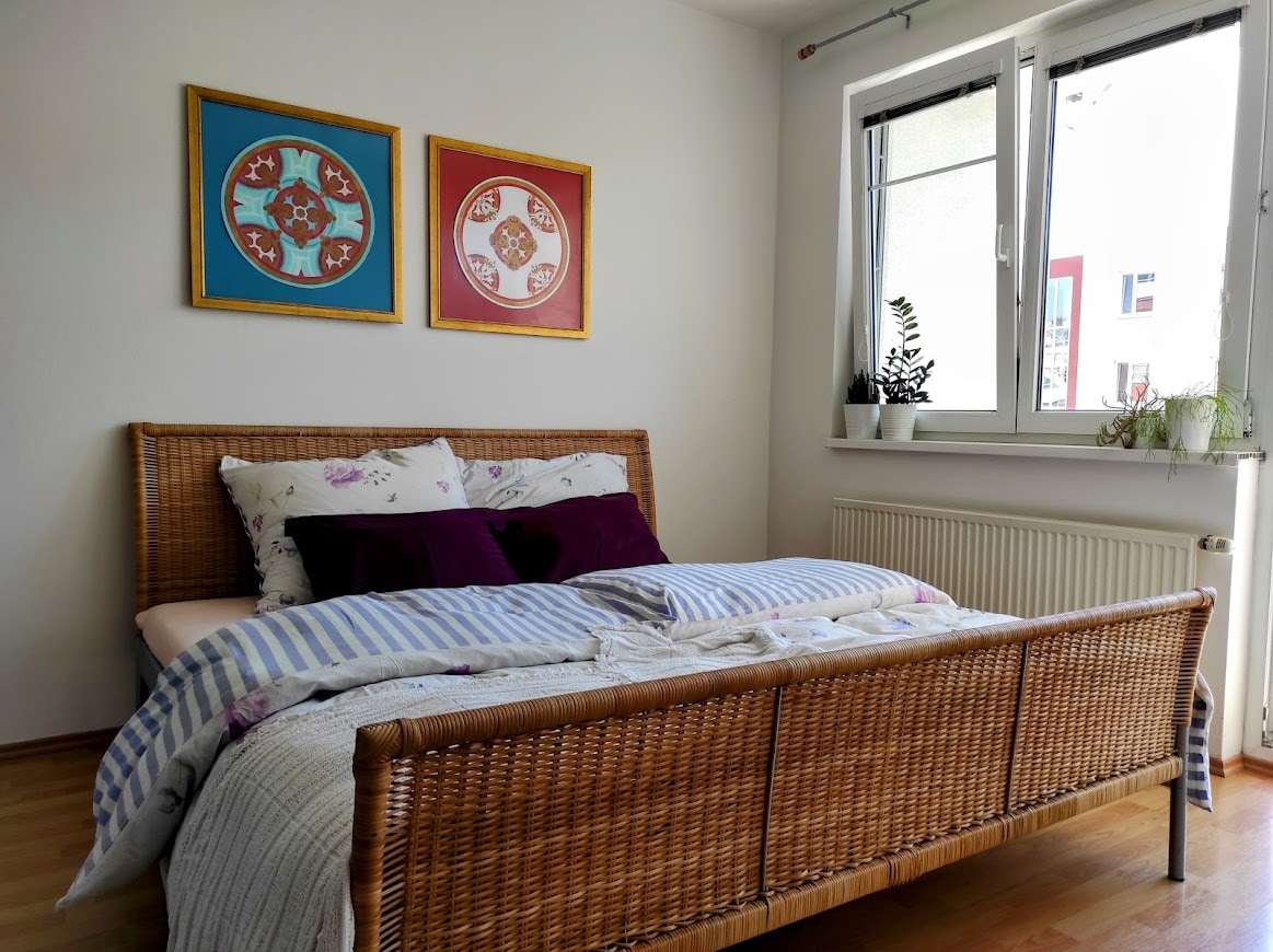 A spacious bedroom features a woven bed frame adorned with soft bedding and pillows in varying shades. Two framed artworks hang above the bed, and a window allows natural light to illuminate the room. A radiator and potted plants contribute to the decor.