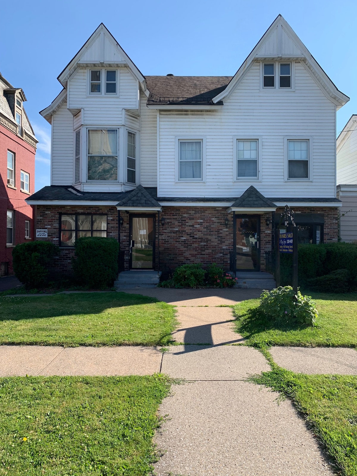 A historic, multi-story building features a blend of brick and white siding. The front yard is well-maintained with grass and bushes, while a paved pathway leads to the entrance. Windows are adorned with light curtains, allowing natural light to fill the interior.