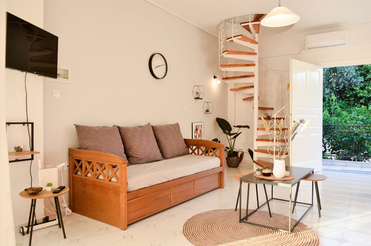 A light-filled living area features a cozy sofa with neutral cushions, a round coffee table, and wall-mounted TV. A decorative clock is displayed on a neutral wall. A staircase leads to the second floor, while a door opens to a balcony with views of greenery.