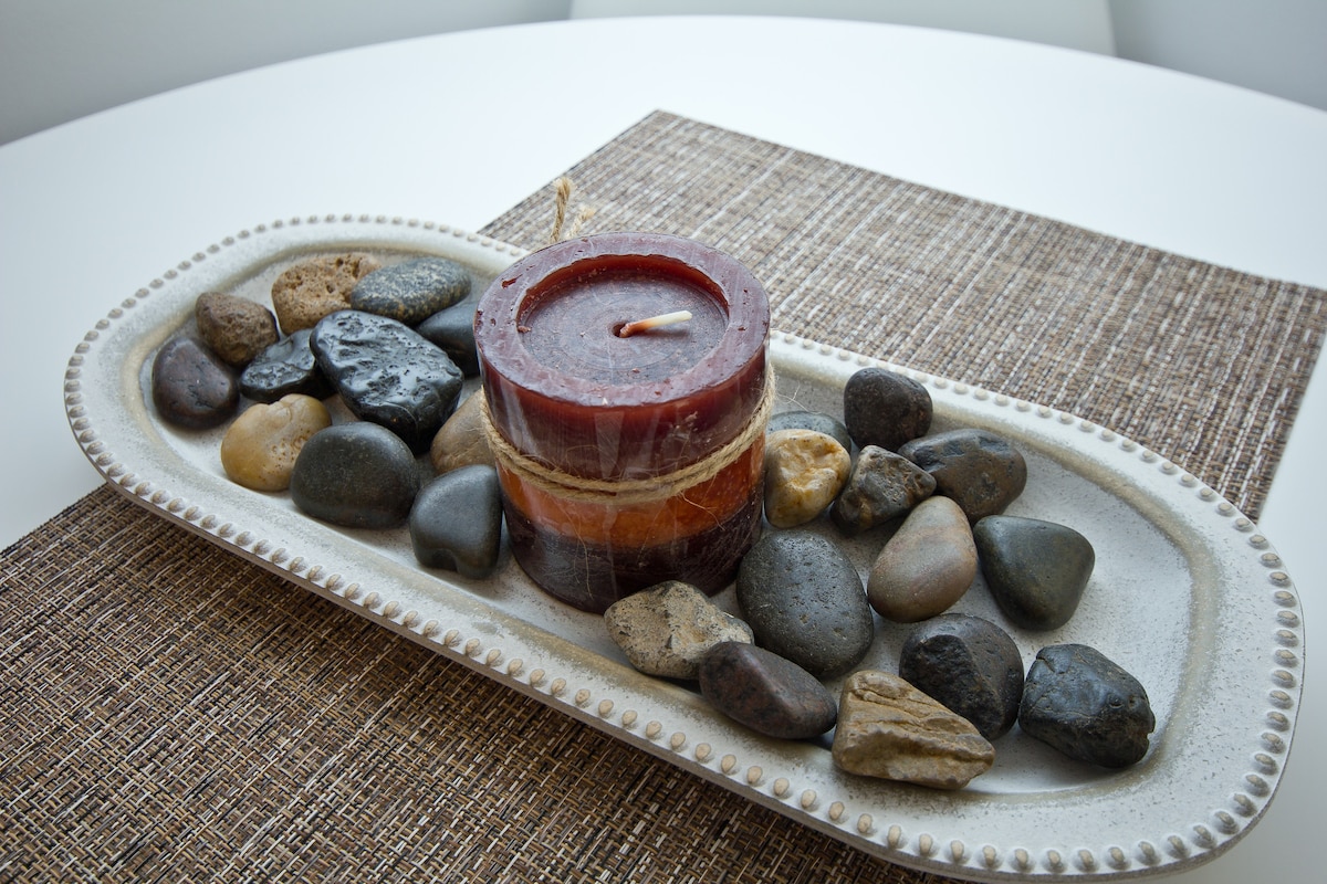 A decorative centerpiece is displayed on a textured table mat, featuring a round candle surrounded by smooth river stones. The candle is layered in shades of deep red and orange, creating a calming focal point within the space.