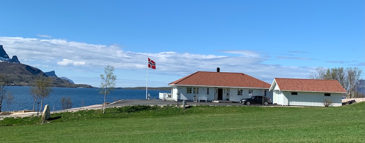 A well-maintained villa is situated along the water's edge, featuring a red-tiled roof and white exterior. Lush green grass surrounds the property, with a Norwegian flag displayed prominently. The serene body of water extends in the background, complemented by a clear blue sky.