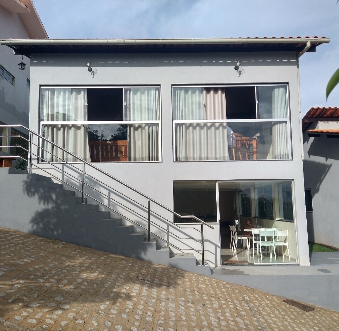 A modern two-story building features large glass windows, allowing natural light to fill the interior. A set of stairs leads up to the covered entryway, with a dining table visible through the glass doors. Light-colored curtains provide a soft texture to the exterior.