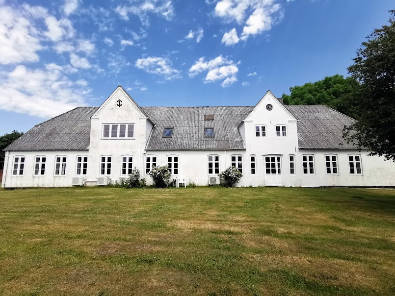 The exterior of a spacious, two-story building is visible, featuring white walls and a sloping grey roof. Multiple windows allow natural light, while the large grassy area in front adds to the property's open feel. Green trees line the background under a partly cloudy sky.