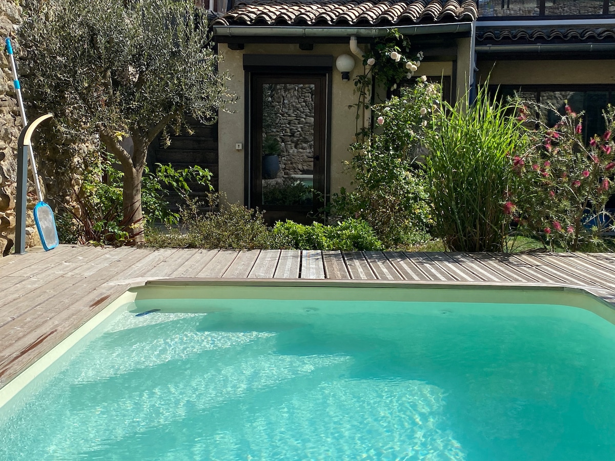The image displays a serene pool surrounded by wooden decking, with lush greenery and blooming plants visible. The pool's clear water reflects the sunlight, while a stone wall features in the background, providing a rustic touch to the outdoor space.