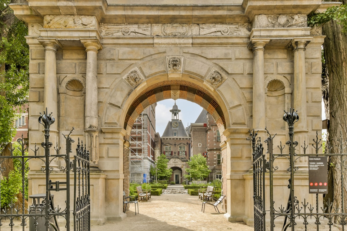 An arched entranceway framed by intricate stonework welcomes visitors, leading to a green courtyard. Benches are positioned along the path, with the historical building visible in the background, featuring a peaked roof and clock tower. Lush trees provide a serene atmosphere.