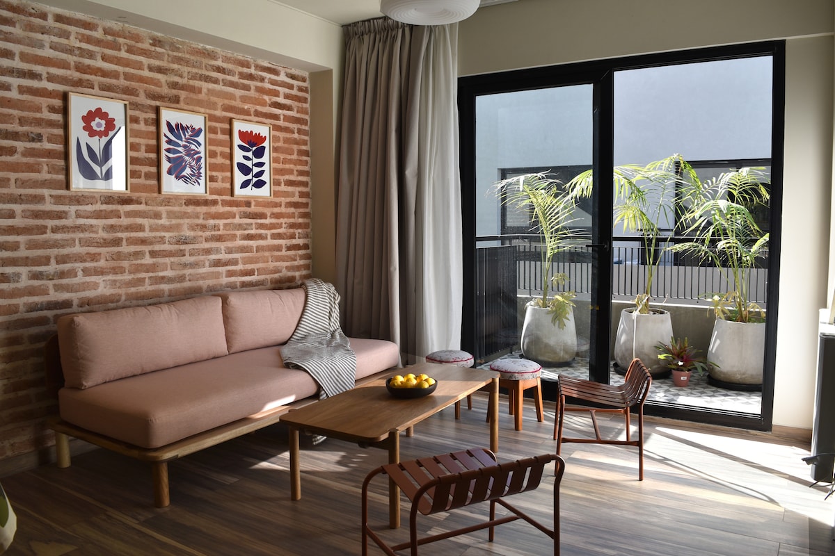 A bright living area features a pink sofa against a textured brick wall, complemented by colorful framed artwork. A wooden coffee table is positioned in front of the sofa, surrounded by stylish seating. Large glass doors open to a balcony decorated with potted plants.