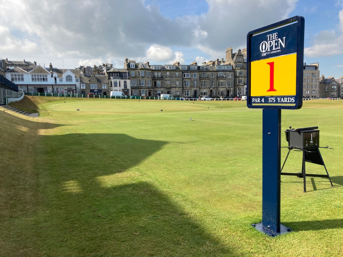 A vibrant sign marks the start of the first hole at The Open, displaying details of the par 4 and its length of 375 yards. A lush green golf course stretches out in the background, accompanied by historical buildings under a clear sky.