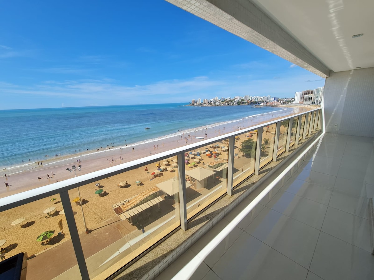 A spacious balcony offers a panoramic view of the beach, showcasing golden sands and azure waters. Colorful beach umbrellas dot the shoreline, while a distant skyline of buildings and people enjoying the beach can be seen under a clear blue sky.