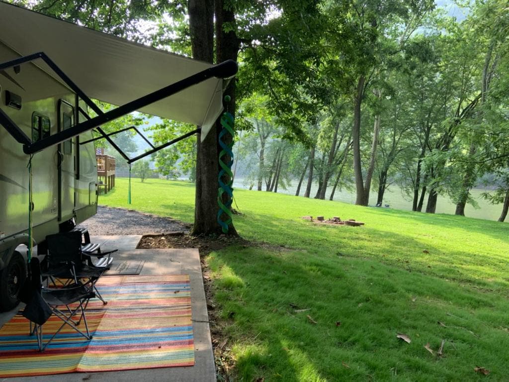 An RV is positioned on a concrete pad, shaded by trees. Two dark folding chairs are set up on a colorful woven rug. A grassy area leads towards a fire pit and the river, with a view of trees lining the water's edge.