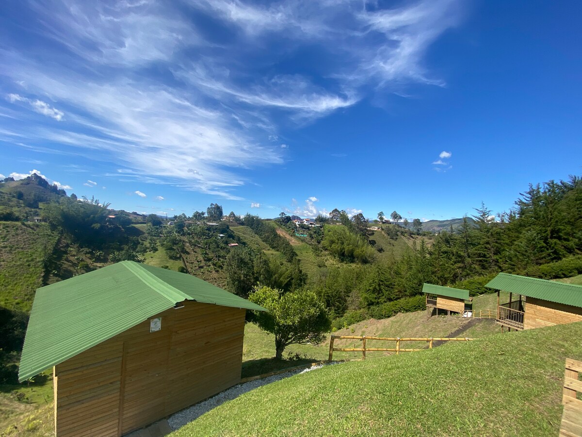 A panoramic view showcases wooden cabins with green roofs set against a backdrop of rolling hills. Lush greenery surrounds the area, with trees dotting the landscape and a clear blue sky overhead. Paths are visible leading through the terrain.