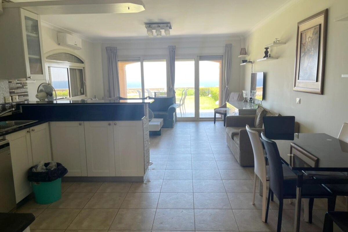 An open living space showcases a modern kitchen with a breakfast bar, leading to a dining area with four chairs. Large glass doors provide a view of the garden and the Red Sea, allowing natural light to fill the room. Neutral tones are complemented by a cozy seating area.