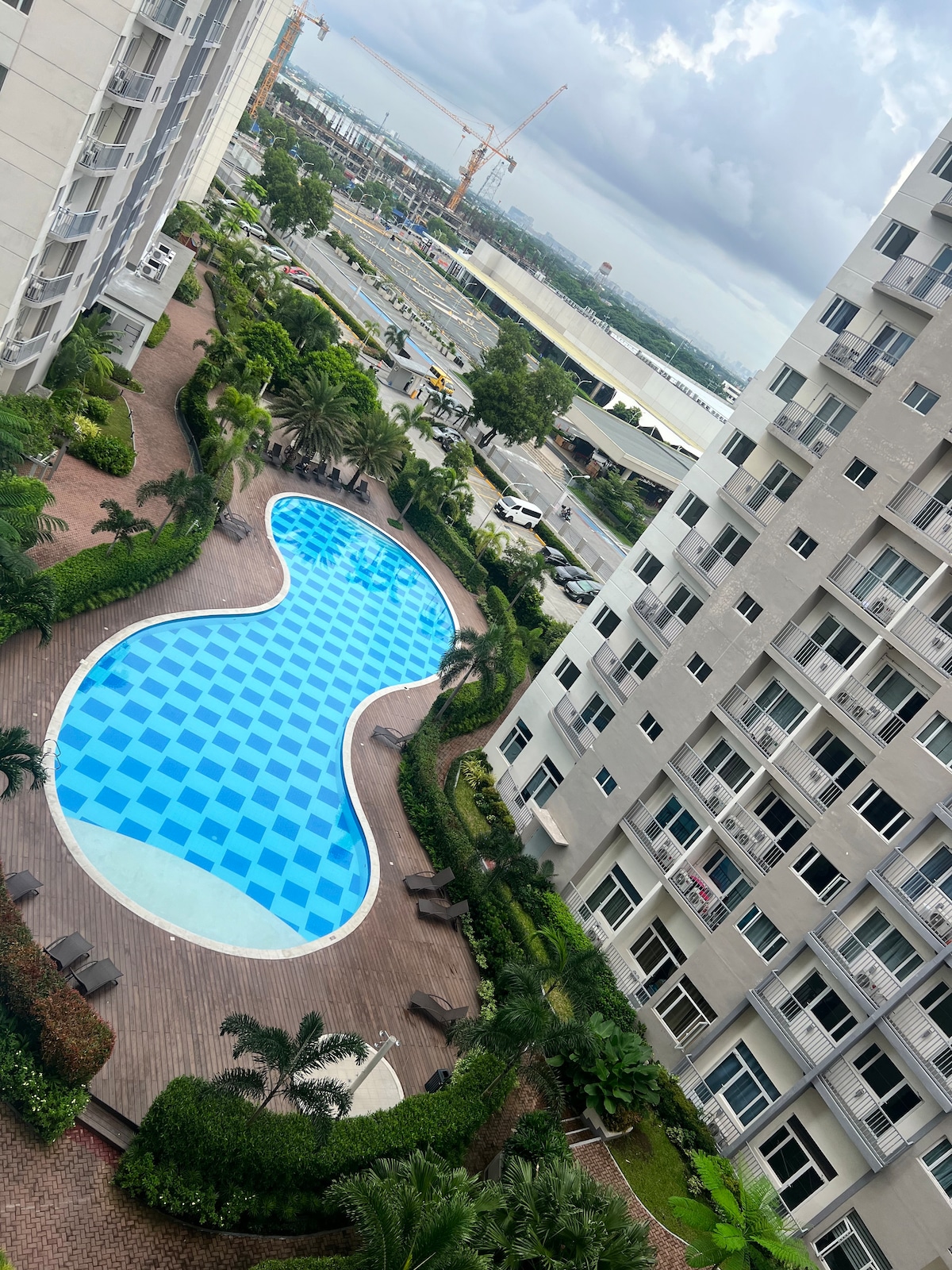 An aerial view of a swimming pool, shaped in a flowing design and adorned with blue and white tiles. Surrounding greenery features palm and other plants, complemented by lounge chairs along the poolside. Nearby, residential buildings are visible, emphasizing a maintained environment.