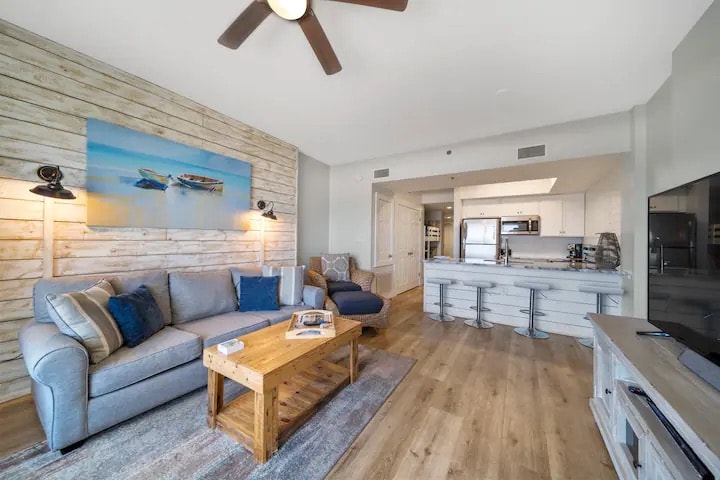 A living area features a comfortable gray sofa and a blue accent chair, positioned around a rustic wooden coffee table. Light wooden paneling adorns the walls, complemented by a coastal-themed artwork. A modern kitchen with a breakfast bar is visible in the background.