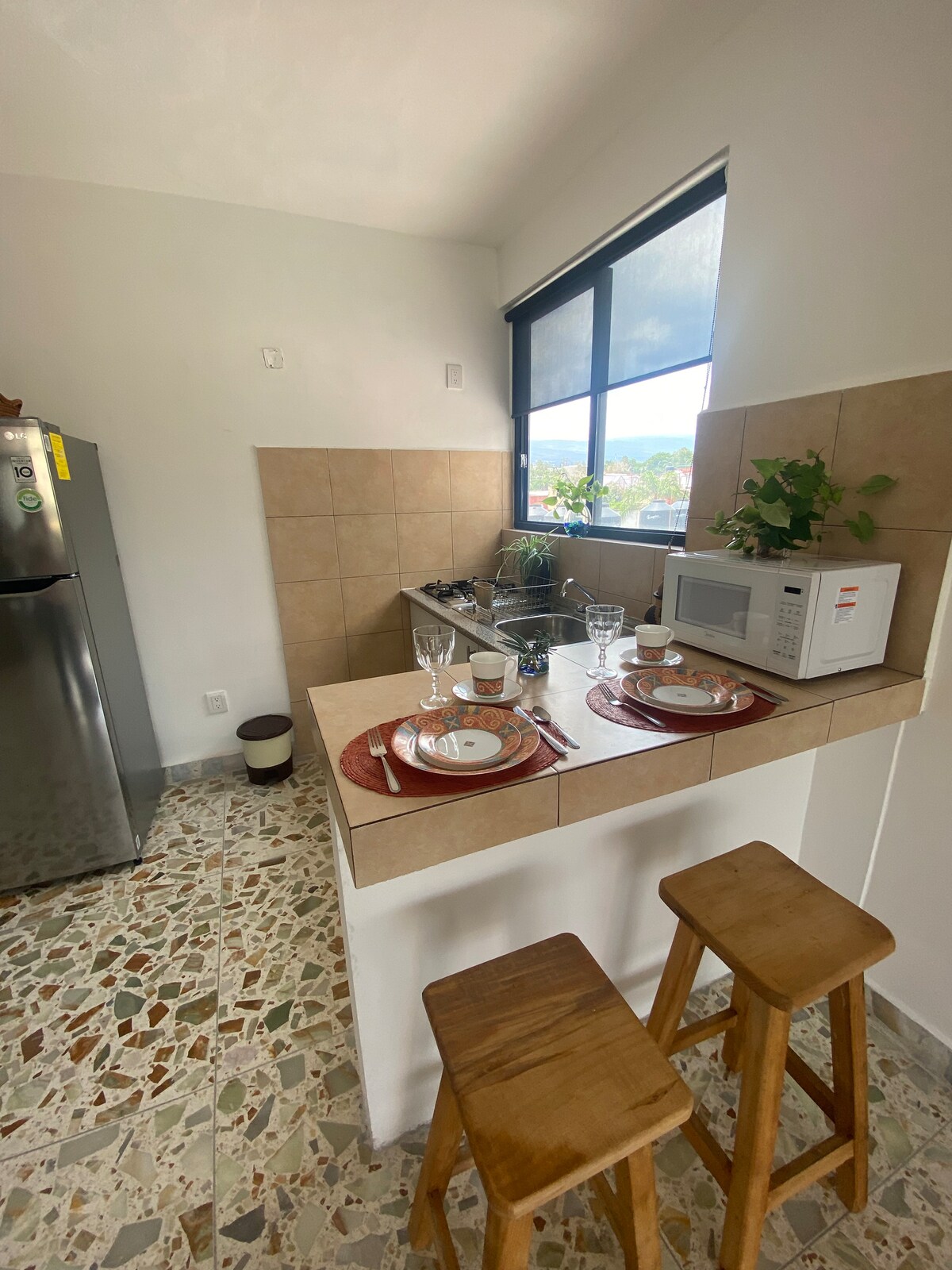 The kitchen features a compact layout with a light-colored tile backsplash. A small bar area with two wooden stools is set for a meal, including two plates and drinkware. A microwave and a refrigerator are visible, with natural light streaming through the window.