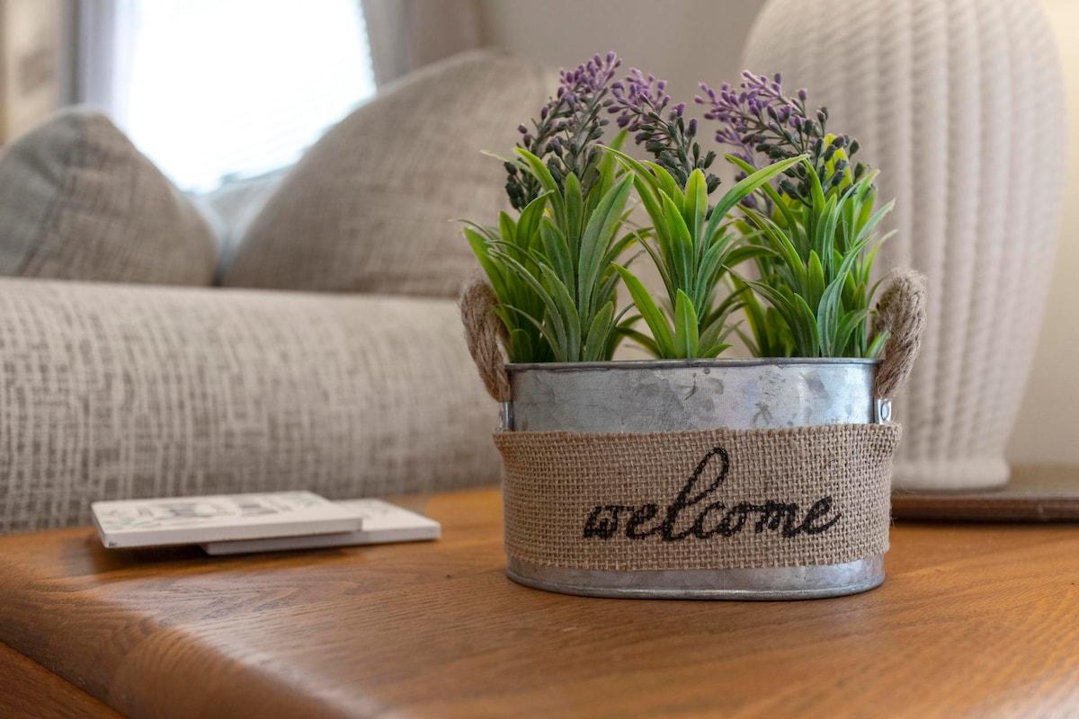A decorative plant arrangement featuring artificial lavender and green leaves sits in a metal container with a burlap band that reads 'welcome.' The container is placed on a wooden table alongside a textured cushion and a softly lit lamp.